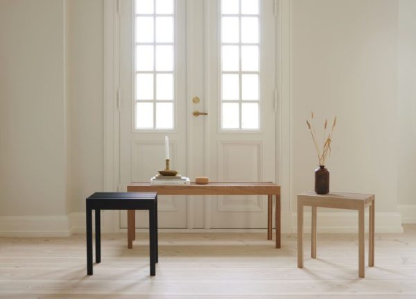 Et minimalistisk værelse med et lyst trægulv med tre FORM & REFINE - Letvægtstaburet i sort, brun og beige, arrangeret asymmetrisk. et stort vindue med hvide døre giver naturligt lys, og en vase med tørrede grene står på det beige skrivebord.