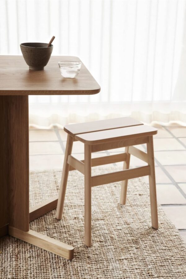 Et minimalistisk værelse med et FORM & REFINE - Quatrefoil Bord 68x68 bord med en keramisk skål og en træskammel nedenunder. Et glas vand står på gulvet, og et rent gardin filtrerer lyset i baggrunden.