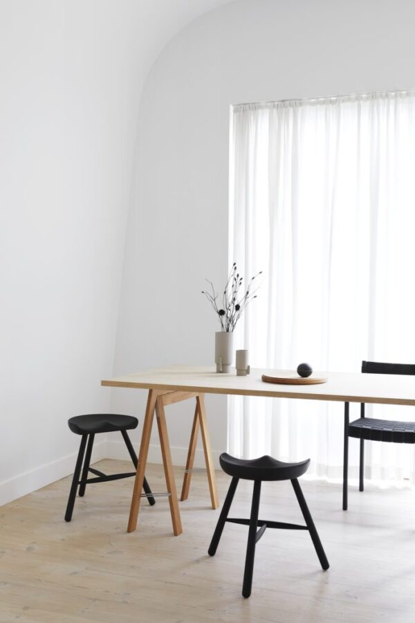 En minimalistisk spiseplads med et FORM & REFINE - Shoemaker Taburet No. 49 bord af naturligt træ, to sorte stole og en bænk. en neutral-tonet vase med grene står på bordet mod en hvid, buet væg og gennemsigtige gardiner.