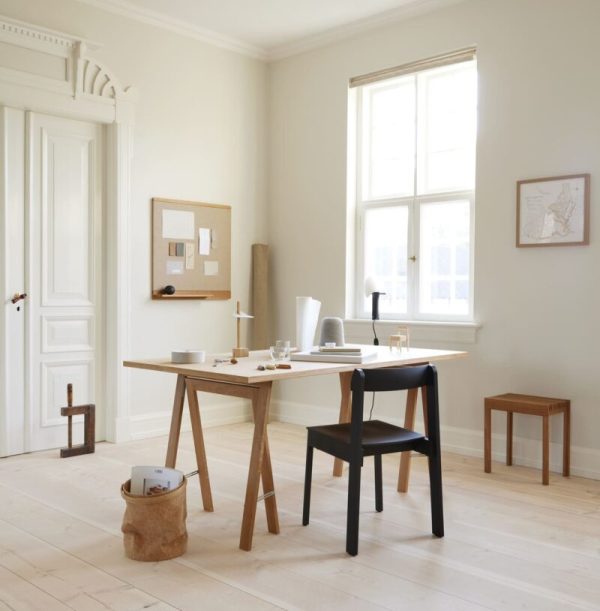 Et minimalistisk hjemmekontor med en FORM & REFINE - Letvægts Taburet og sort stol, sat i et lyst rum med beige vægge, to vinduer og et lyst trægulv. en korktavle, et kort og en bærbar computer dekorerer rummet.