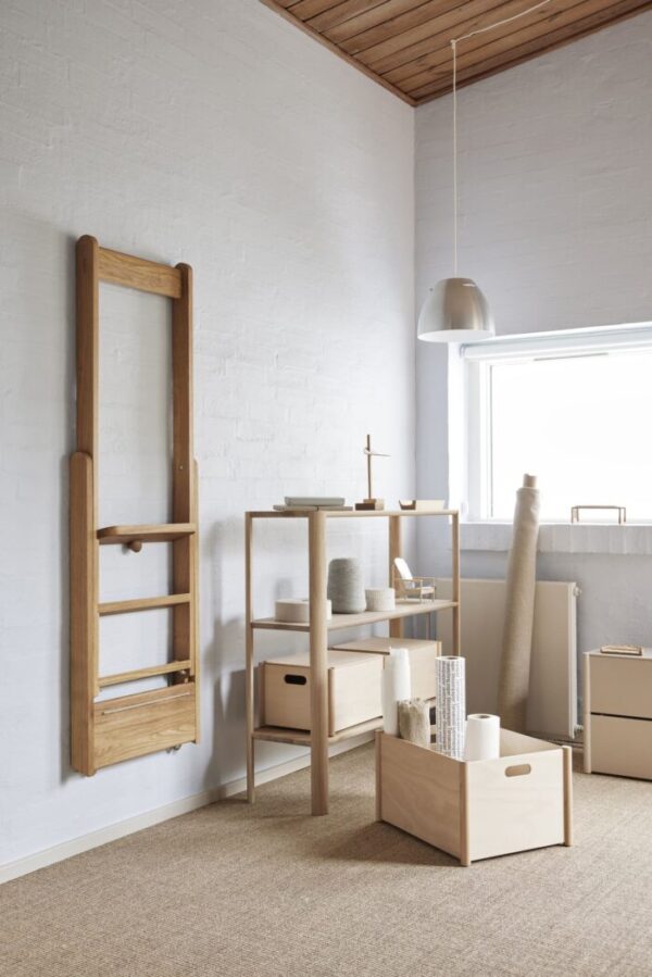 Et minimalistisk værelse med hvide murstensvægge med en træstige og en opbevaringshylde med forskellige ting. FORM & REFINE - Leaf Reol 1x3 er på gulvet nær et stort vindue.