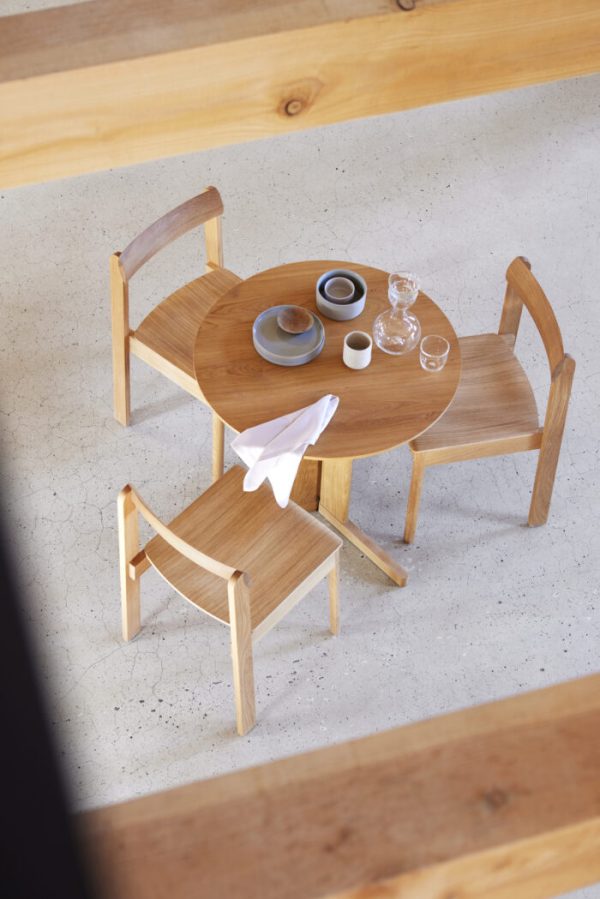 En minimalistisk spisestue med to træstole og en FORM & REFINE - Trefoil Bord Ø75. på bordet står et par fade, kopper og en spildt serviet, alt sammen under naturlig belysning.
