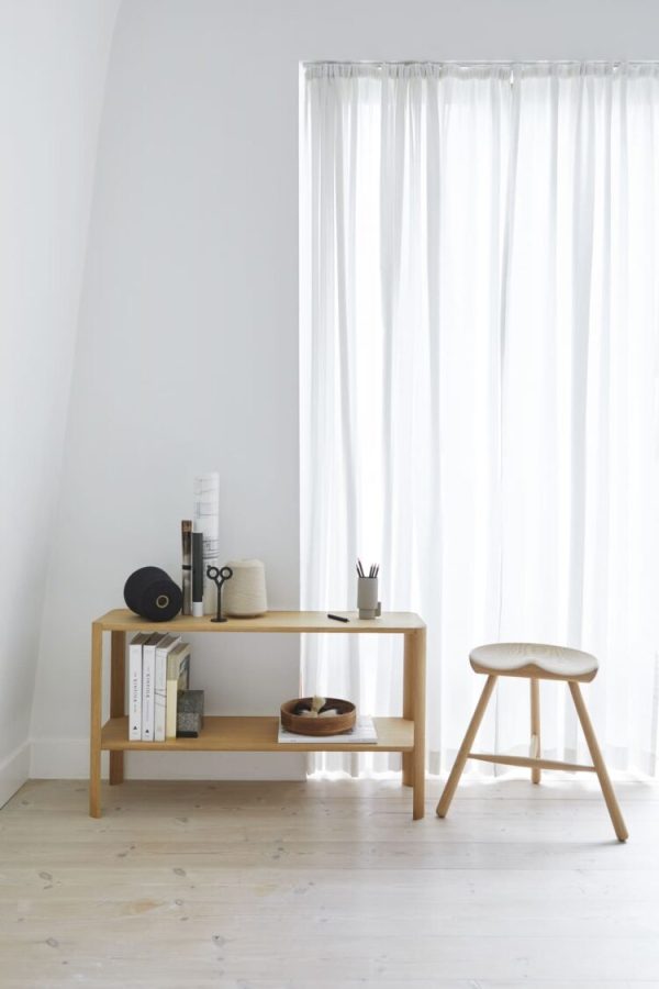 Et minimalistisk værelse med hvide vægge og et trægulv med et Leaf Reol 1x2 konsolbord med bøger, en skål og papirvarer. en lille skammel er placeret til højre, nær et vindue med rene hvide gardiner.