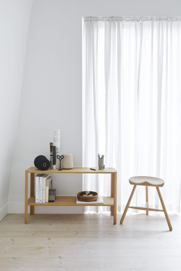 Et minimalistisk setup med en FORM & REFINE - Shoemaker Taburet nr. 49 med bøger, dekorationsgenstande og en stofrulle ved siden af en lille skammel. rummet har hvide vægge, gennemsigtige gardiner og lyse trægulve.