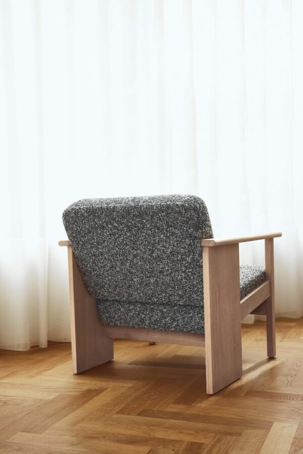 En moderne stol med træramme og tekstureret gråt polstring, stående på et sildebensparketgulv mod en baggrund af rene hvide gardiner. Dette stykke afspejler exceptionelt designhåndværk.