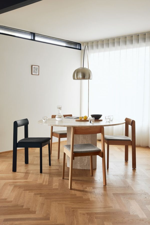 En moderne spisestue med et træbord sat med tallerkener og glas, omgivet af tre FORM & REFINE - Blueprint Stol Hallingdal stole i forskellige stilarter, under en stor pendel, med et lyst vindue og et lille indrammet billede på væggen.