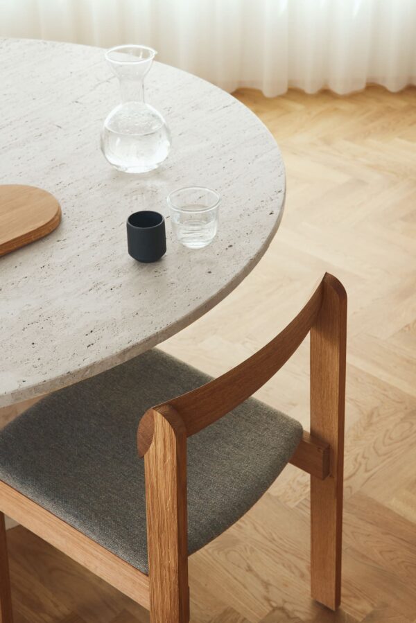 En minimalistisk spisestue med et rundt terrazzobord med en lille sort kop og to glaskarafler. A FORM & REFINE Blueprint Stol Hallingdal med gråt polstret sæde er gemt under bordet, sat på et lyst trægulv.