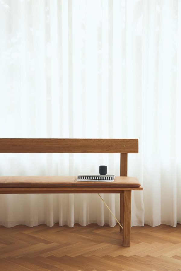 Et minimalistisk interiør med en FORM & REFINE - Placer Bænk 155 mod en væg med rene hvide gardiner. på bænken er der en lille sort højttaler og en notesbog med en pen. blødt naturligt lys oplyser scenen.
