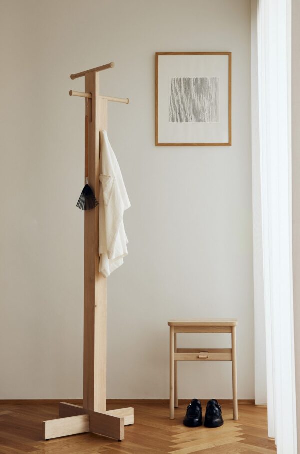 Et minimalistisk værelse med en knageholder i træ med et hvidt håndklæde og en kvast ornament. Et indrammet abstrakt kunstværk fra TomorrowsDesign hænger på væggen. En træskammel og sorte sko er placeret i nærheden på gulvet.