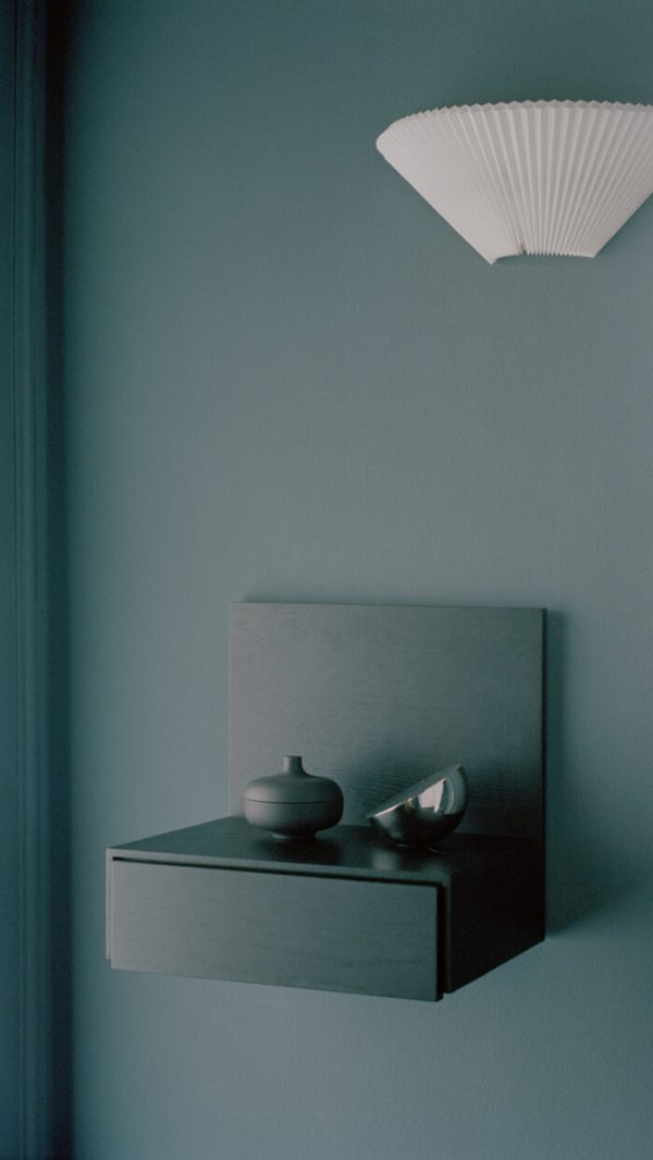 Et minimalistisk interiør med et NYT VÆRK - Tana Væghængt Natbord på en blå væg med en keramisk gryde, en metallisk skål og en dekorativ væglampet formet som en hvid vifte over.