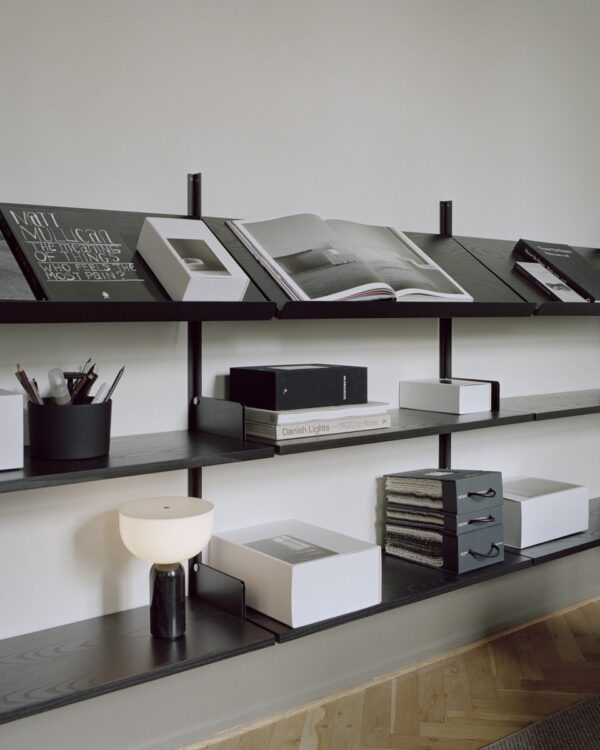 Fire parallelle hylder på en beige væg med forskellige ting: åbne bøger, æsker, en lampe og stationære holdere i et rent og moderne TomorrowsDesign-arrangement.