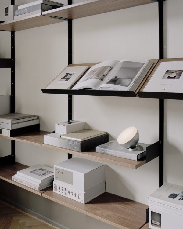 En minimalistisk møbler-reol med åbentstående kunstbøger, dekorative genstande og pænt stablede kasser. En lille lampe er placeret på en af hylderne, som giver et varmt skær til omgivelserne.