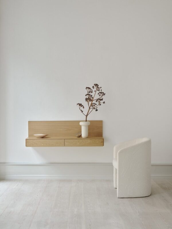 Et minimalistisk rum med et NYT VÆRK - Tana Væghængt Mediemodul monteret på en lysegrå væg med en vase med tørrede blomster. ved siden af står en cremefarvet tekstureret lænestol på et lyst trægulv.
