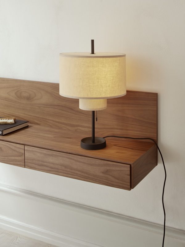 ET NYT VÆRK - Tana Væghængt Mediemodul monteret på en lys beige væg med en mørkebrun bordlampe med cylindrisk skærm. modulet rummer også en bog og børste, med en stikkontakt synlig nedenfor.