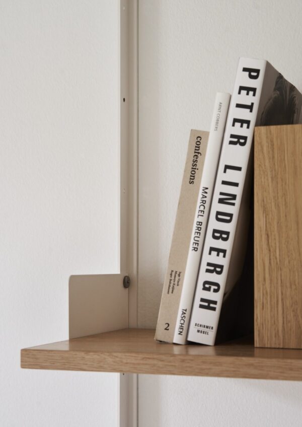 Et hvidt metalbeslag støtter en træhylde mod en hvid væg. Reolen rummer to store bøger med titlen "Peter Lindbergh" og "Conversations on Conflict Photography." Dette minimalistiske design eksemplificerer TomorrowsDesigns etos.