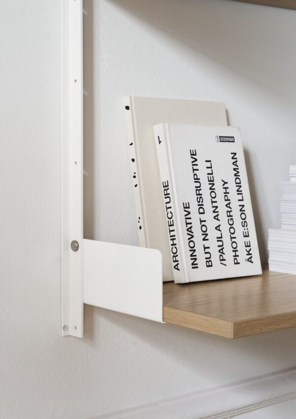 En minimalistisk hylde på en hvid væg med et pænt stablet udvalg af hardcover bøger om arkitektur og designmøbler, med synlige fremtrædende titler.