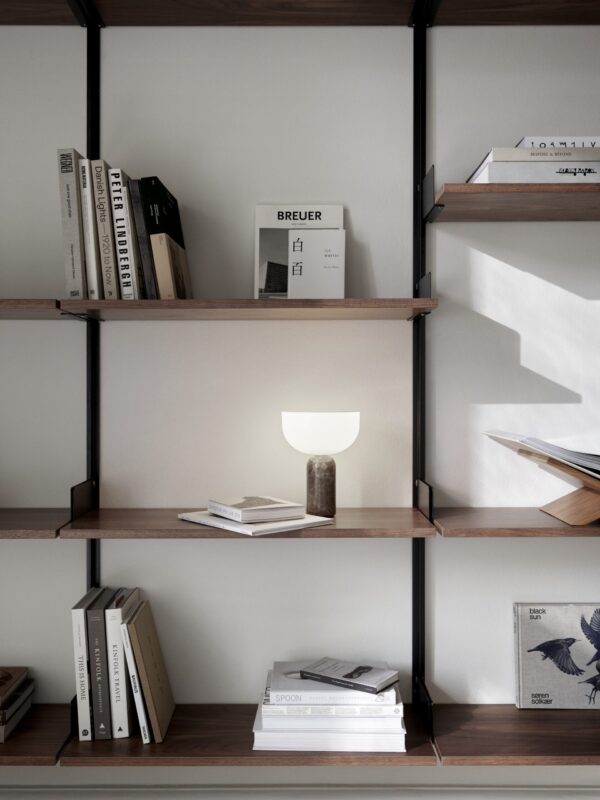 En moderne bogreol med pænt organiserede bøger og pynteting. Hylderne er lavet af træ og understøttet af en metalramme, med en lille, oplyst lampe og forskellige bøger om arkitektur og TomorrowsDesign.
