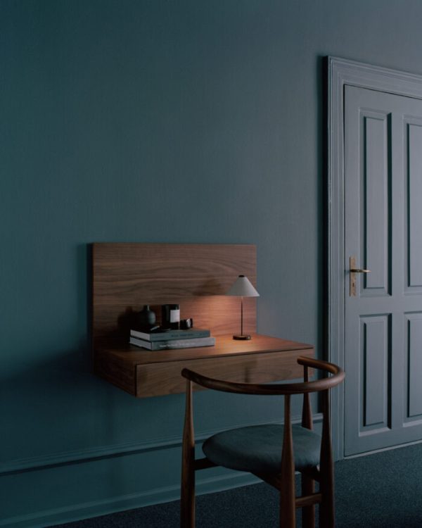 Et godt oplyst rum med et NYT VÆRK - Tana Væghængt Skrivebord, en stol og en lampe. bøger er pænt arrangeret på skrivebordet, og en lukket hvid låge er til højre. den overordnede stemning er rolig og minimalistisk.