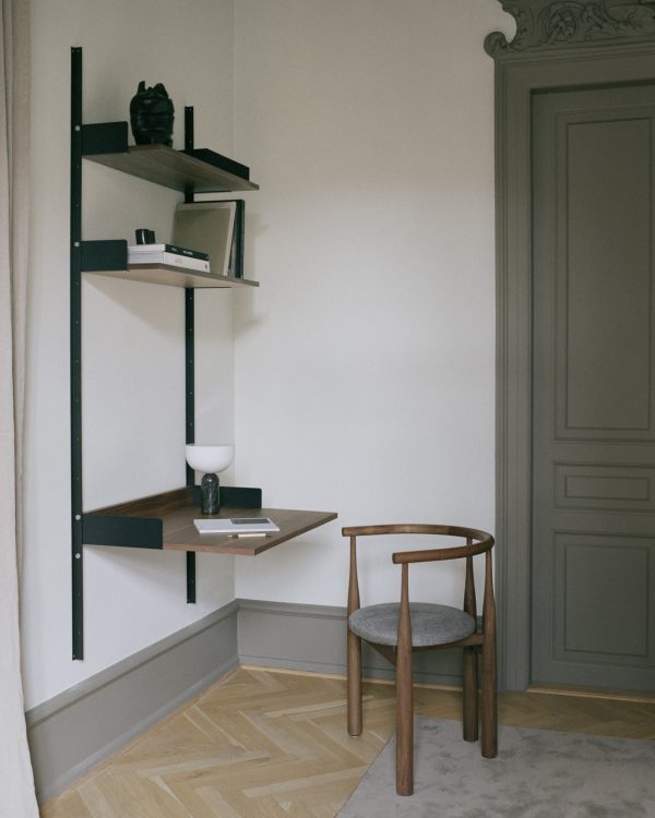 Et minimalistisk hjørne af et værelse med en træstol og modulopbygget reol med bøger, en lampe og dekorative genstande. Rummet har et sildebensgulv og en detaljeret grå dørkarm fra TomorrowsDesign.