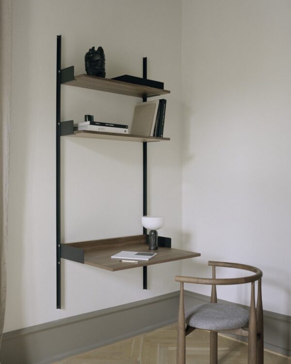 Et minimalistisk værelseshjørne med en lodret vægmonteret hylde med tre niveauer med bøger, en lille skulptur og dekorative genstande ved siden af en enkel møbleret træstol med en grå hynde.