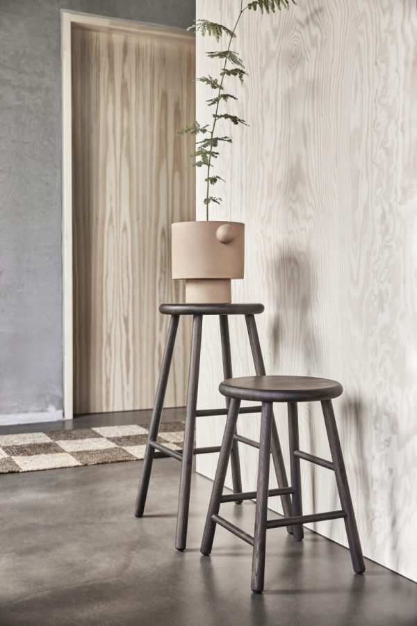 Et minimalistisk interiør med to OYOY - Moto Stol Lav, en højere med en potteplante på toppen, mod en tekstureret trævæg og betongulv. et mønstret tæppe er delvist synligt.