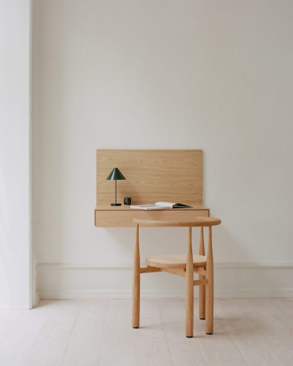 ET NYT VÆRK - Tana Væghængt Skrivebord og bænk mod en lys beige væg i et stærkt oplyst rum. skrivebordet har en lille grøn lampe, en åben bog og et par briller på.