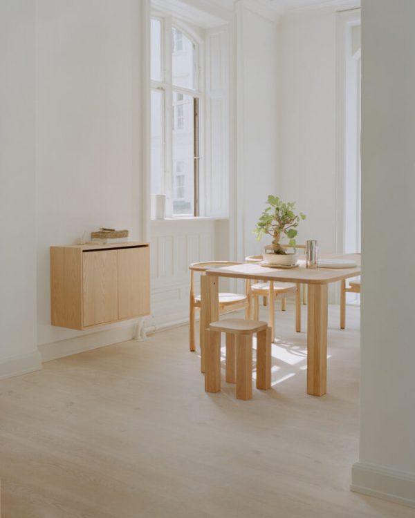 Et minimalistisk værelse med trægulv og et enkelt bord og taburetter. ET NYT VÆRK - Vægskab Høj M. Døre skab og en plante på bordet er ved et stort vindue, der oversvømmer rummet med naturligt lys.
