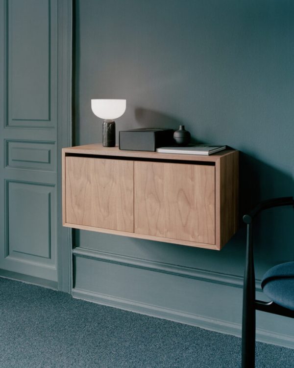 En minimalistisk gang med et NYVÆRK - Vægskab Lav M. Døre mod en blågrå væg. skabet rummer en hvid lampe og sorte bøger. der er også en sort stol delvist synlig til højre.