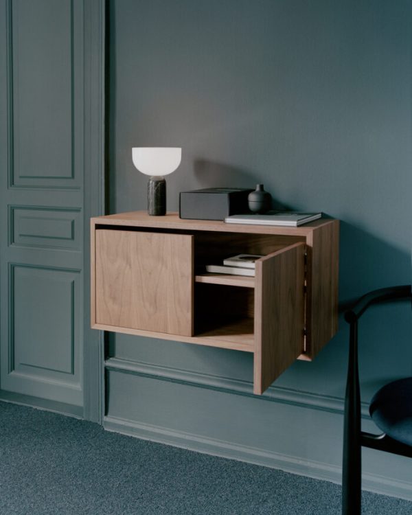 Et minimalistisk NYT VÆRK - Vægskab Low M. Døre mod en grå væg, med åbent skab og en lampe på toppen, ved siden af en sort stol og et blågrønt gulvtæppe.