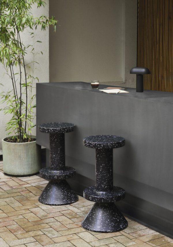 En elegant udendørs indstilling med to NORMANN COPENHAGEN - Bit Barstol 75 taburetter ved siden af en slank mørkegrå bordplade, suppleret med en grøn potteplante og en kop på overfladen.