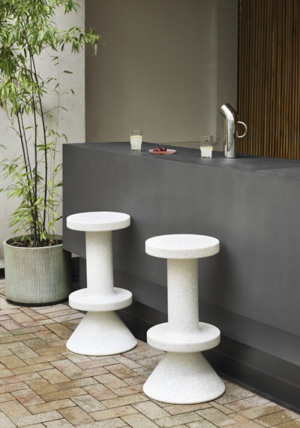 Udendørs terrasse med en slank, moderne NORMANN COPENHAGEN - Bit Barstol 75 med tre cylindriske skamler og en potteplante. To glas og en skål kirsebær stilles på barens overflade.