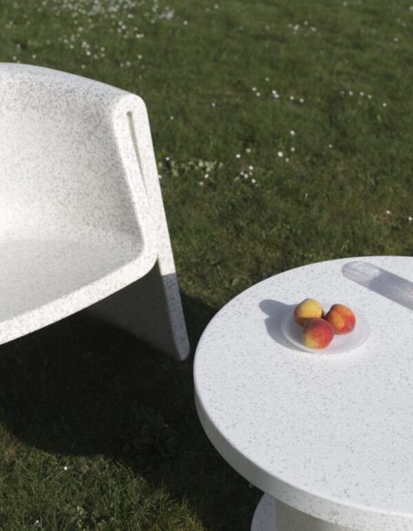 En moderne, tekstureret hvid NORMANN COPENHAGEN - Bit Loungestol og et matchende cirkulært bord sidder på en græsplæne drysset med små hvide blomster. tre modne ferskner placeres på en hvid tallerken oven på bordet.
