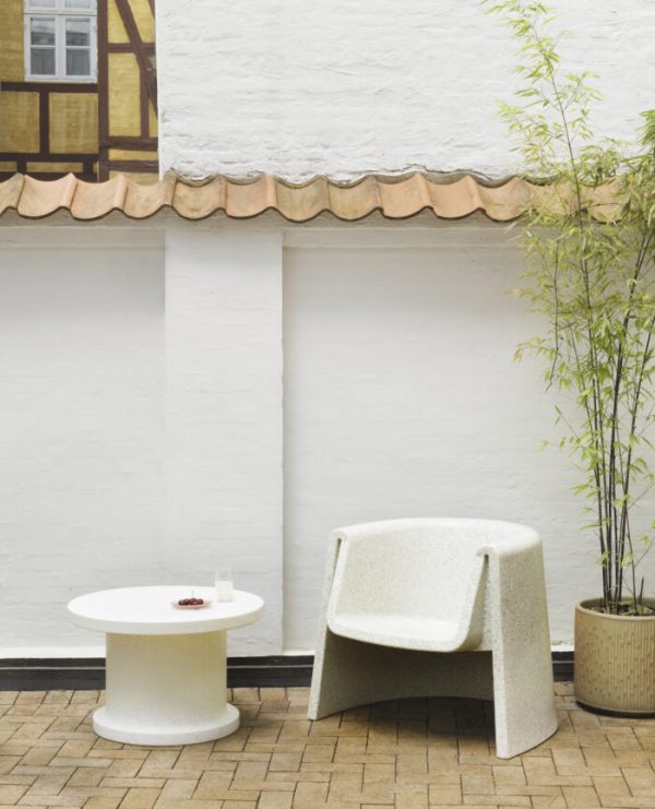 Et roligt gårdhaveområde med en NORMANN COPENHAGEN - Bit Loungestol og et rundt bord med en lille plante på toppen, på baggrund af en hvid væg. En høj bambusplante er i et hjørne ved siden af en traditionel bindingsværksbygning.