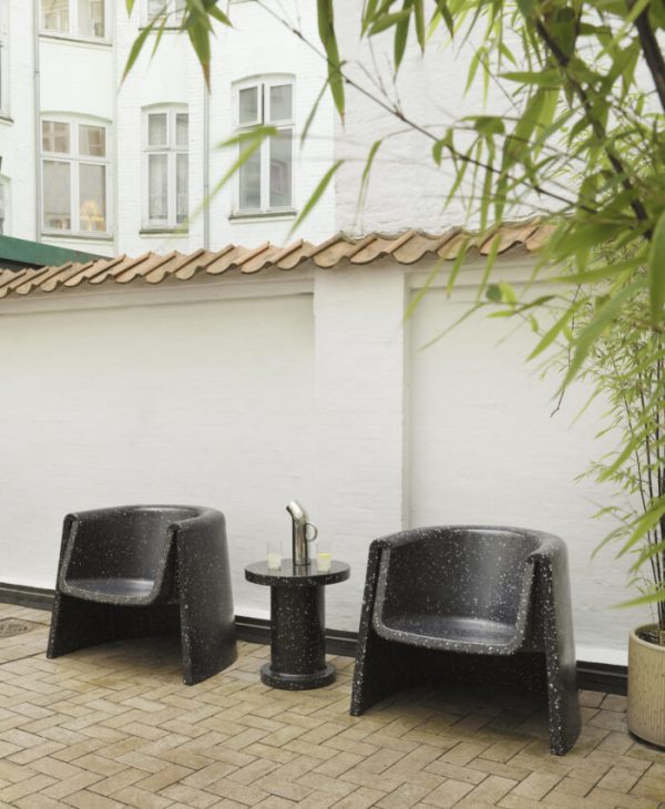 To NORMANN COPENHAGEN - Bit loungestol lænestole og et lille rundt bord med en kaffekop og bog, placeret på en beige fliseterrasse. en hvid væg og grønt løv i baggrunden supplerer de fredfyldte udendørs omgivelser.