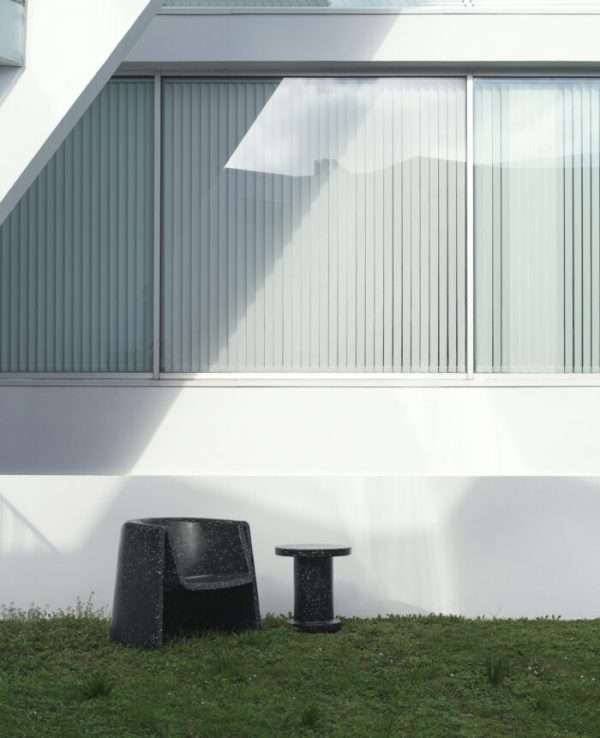 Dette billede viser en moderne bygnings ydre med et stort vindue og hvid facade, en NORMANN COPENHAGEN - Bit Loungestol og matchende skammel er placeret på græs i forgrunden i skyggen af bygningen.