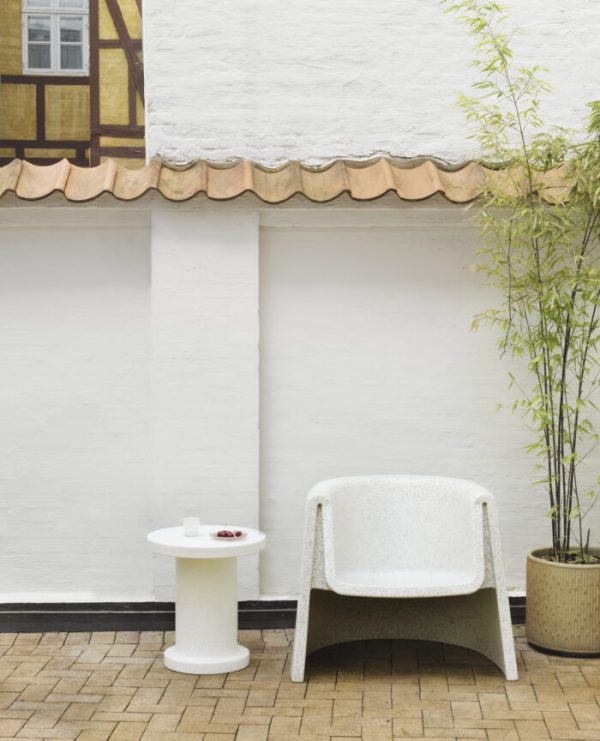 Et moderne udendørs opholdsområde med en hvid lænestol og et lille rundt bord, på en muret terrasse ved siden af en hvid væg med høje tudor-stil overfacader. A NORMANN COPENHAGEN - Bit Sidebord er til højre.