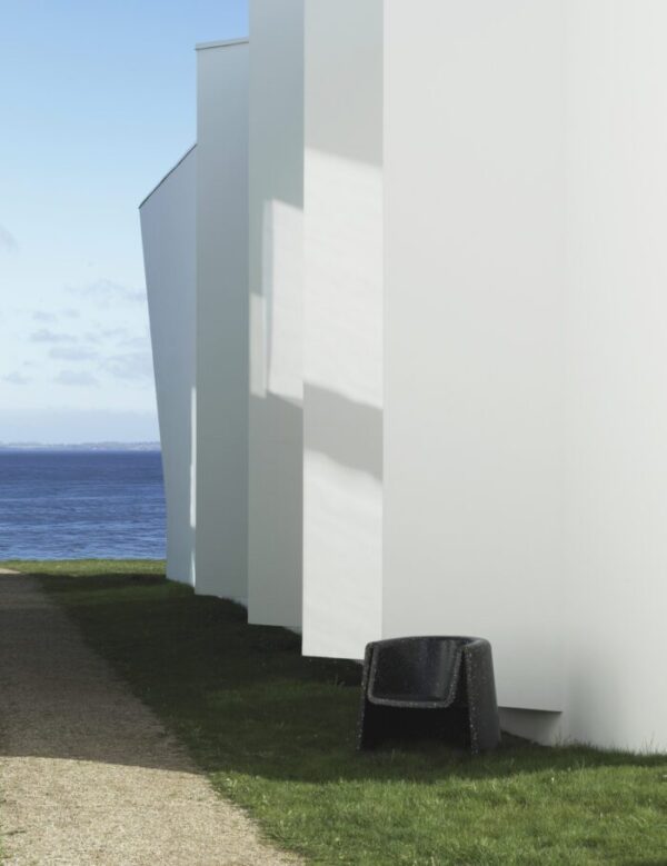 En moderne, kantet hvid NORMANN COPENHAGEN - Bit Loungestol ved siden af en velplejet græsplæne med udsigt over et vandområde med en klar blå himmel over og en sort bænk i forgrunden.