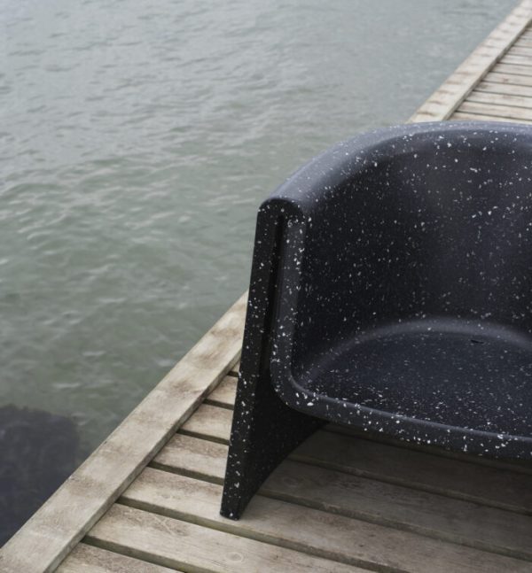 En sort, plastik NORMANN COPENHAGEN - Bit Loungestol med synlige hvide pletter sidder på en træmole, delvist ud over kanten, nær roligt vand. Fokus er på teksturen og detaljerne i NORMANN COPENHAGEN - Bit Loungestol og træet.