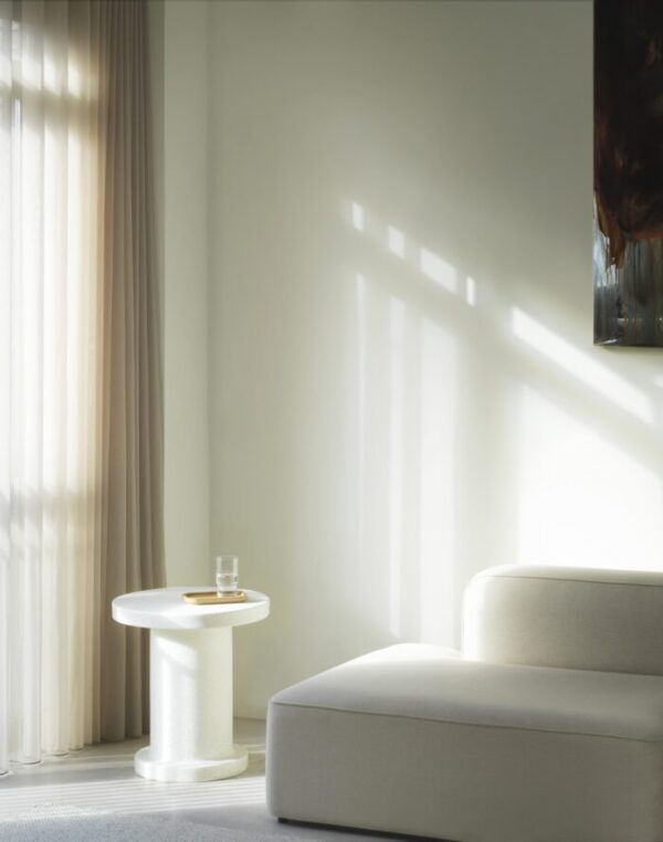 En minimalistisk stue badet i blødt naturligt lys fra et vindue med gennemsigtige gardiner. En moderne hvid sofa og en NORMANN COPENHAGEN - Bit Sidebord med et glas vand på. Sollys kaster geometriske skygger på væggen.