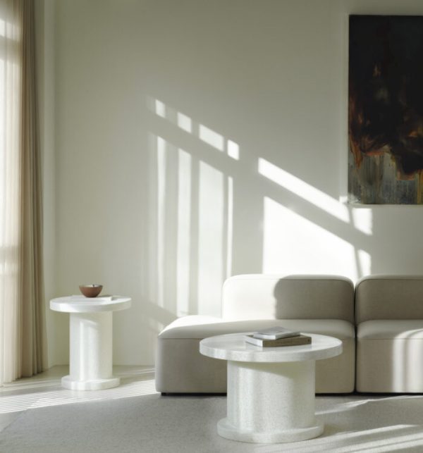 En minimalistisk stue med et NORMANN COPENHAGEN - Bit Sofabord, to moderne hvide borde og et skyggemønster fra vinduesbjælkerne støbt på en væg med subtile kunstværker. rummet er badet i naturligt lys.
