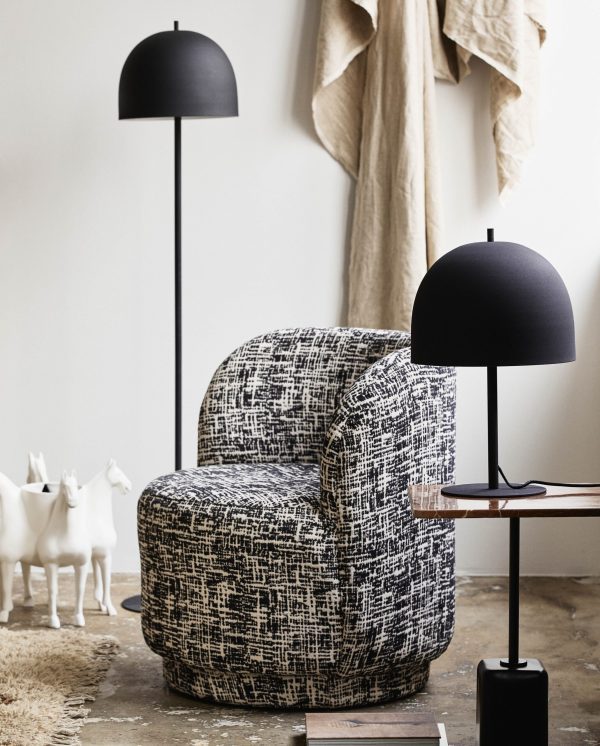 En hyggelig lænestol betrukket med sort og hvidt stof, flankeret af to sorte kuppelgulvlamper, med et sidebord af træ, et draperet beige stof og hvide hestefigurer i baggrunden.