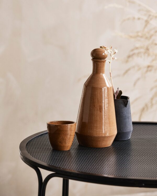 Et keramisk sakesæt med en flaske og en kop i jordfarver og en lille mørk vase med hvide blomster, er udstillet på et sort metalbord mod en tekstureret beige væg.