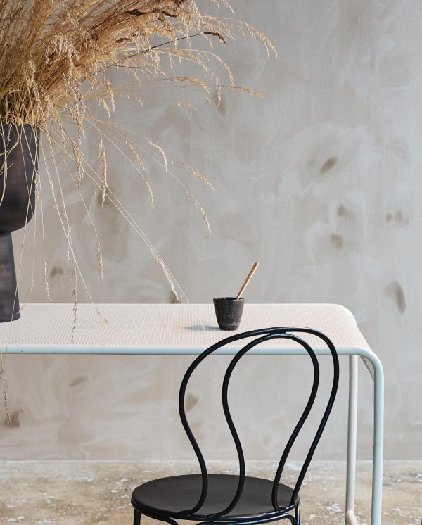 Et minimalistisk interiør med et lyseblåt bord med en skulpturel sort base, parret med en lille sort skammel. en rustik vase med tørrede planter står på bordet mod en tekstureret beige væg.