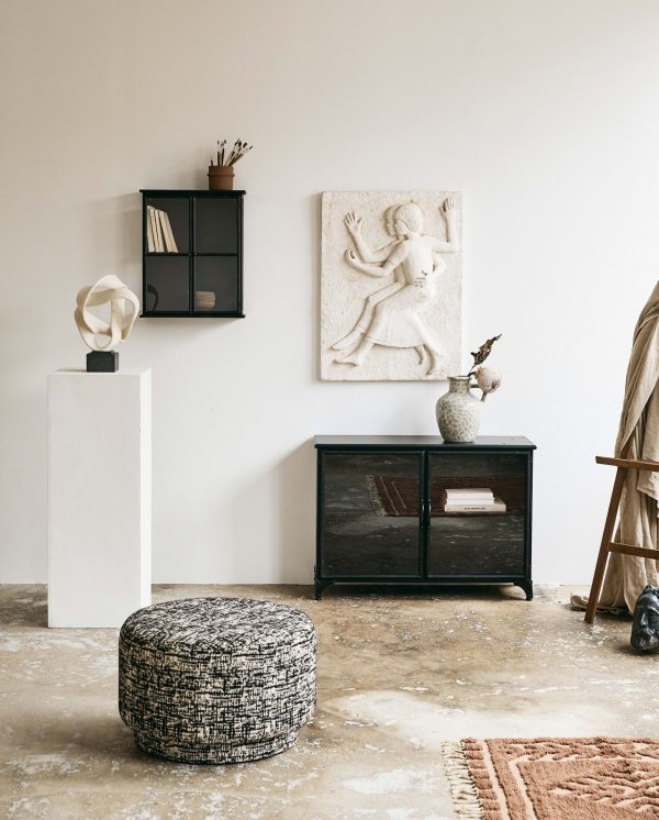 Et minimalistisk værelse med neutrale vægge med en klassisk reliefskulptur, et sort træskab, en hvid skulptur på en piedestal og en mønstret skammel på et tekstureret tæppe.