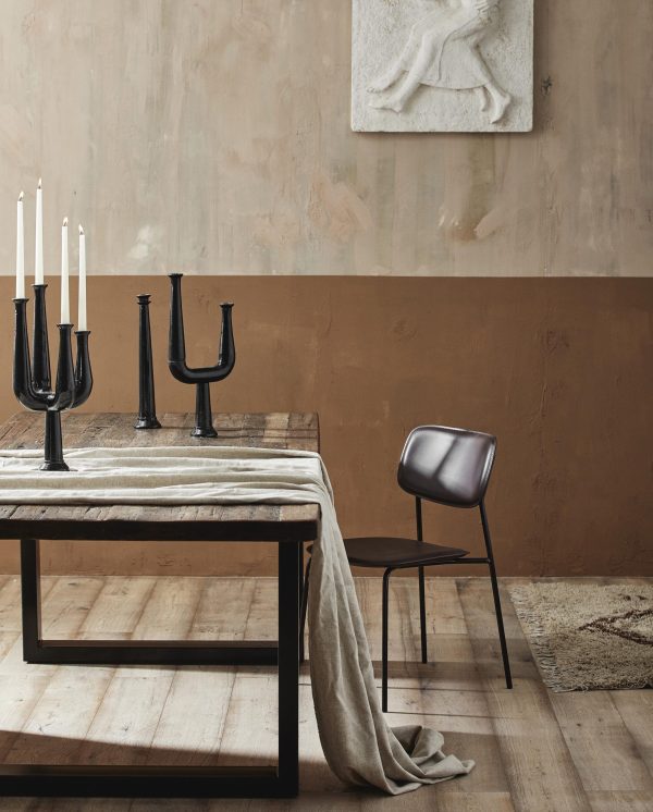 En minimalistisk spisestue med et stenmønstret bord dækket med en beige klud, parret med en slank sort stol. på bordet står høje sorte lysestager. et rustikt tæppe og en basrelief-skulptur pryder rummet.