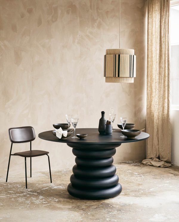 En moderne spisestue med et rundt sort bord med en skulpturel base, parret med en enkel sort stol. bordet er dækket med minimalistisk servise og glas, under en stilfuld hængelampe, mod en struktureret beige væg.