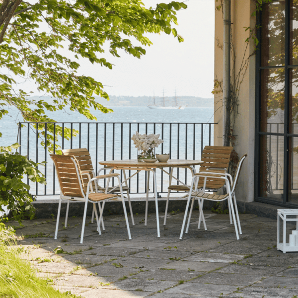 En udendørs terrasse har MANDALAY – Magnolia & Bellevue Havesæt Ø110 med en hvid potteplante på toppen, omgivet af fire træstole med metalstel. Terrassen har udsigt over et vandområde og er omkranset af et sort metalrækværk. Træer giver grønt og skygge.