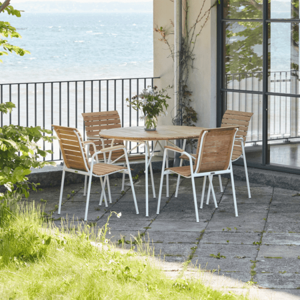 A MANDALAY – Magnolia & Bellevue Havesæt Ø110 med træbord og fire træstole er indrettet på en stenterrasse. En vase med blomster står på bordet. Terrassen ligger ved siden af en glasdør og har udsigt over et vandområde. Grønt løv indrammer scenen.