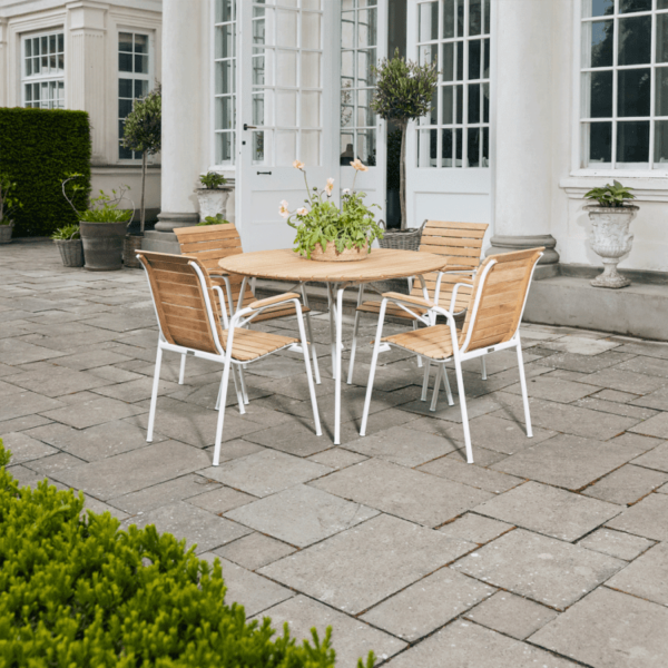 A MANDALAY – Magnolia & Bellevue Havesæt Ø110 med et rundt træbord og fire matchende træstole med hvide metalstel ligger på en stenterrasse. Potteplanter er placeret rundt om terrassen, som ligger foran en hvid bygning med store vinduer og døre.