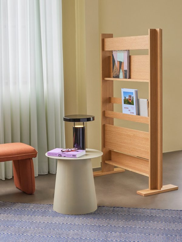 Et moderne interiørdesign med en bogreol i træ med bøger og en billedramme, et rundt hvidt og sort sidebord med bøger på toppen og en ferskenfarvet taburet på et blåt tekstureret tæppe.